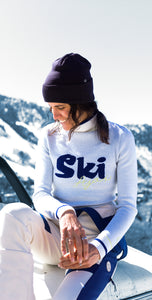 Person wearing a white ski jacket with 'Ski' printed on it, sitting on a car with a snowy landscape in the background.