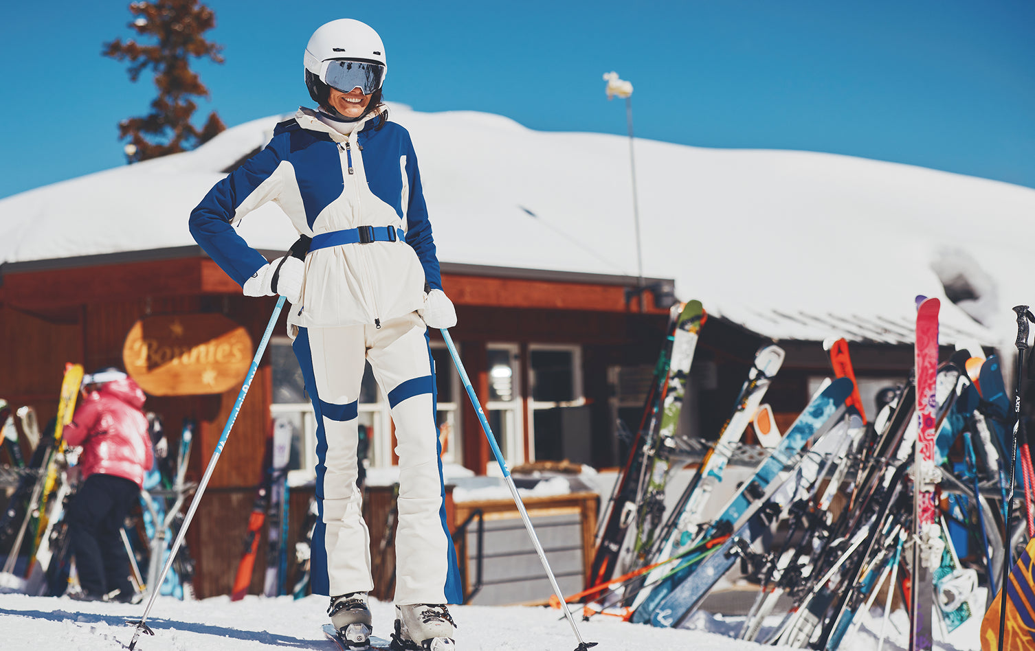 Person in ski gear standing in a snowy area with skis and a building in the background