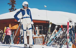 Person in ski gear standing in a snowy area with skis and a building in the background
