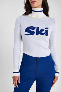 Person wearing a white sweater with 'Ski' text and blue pants on a plain background