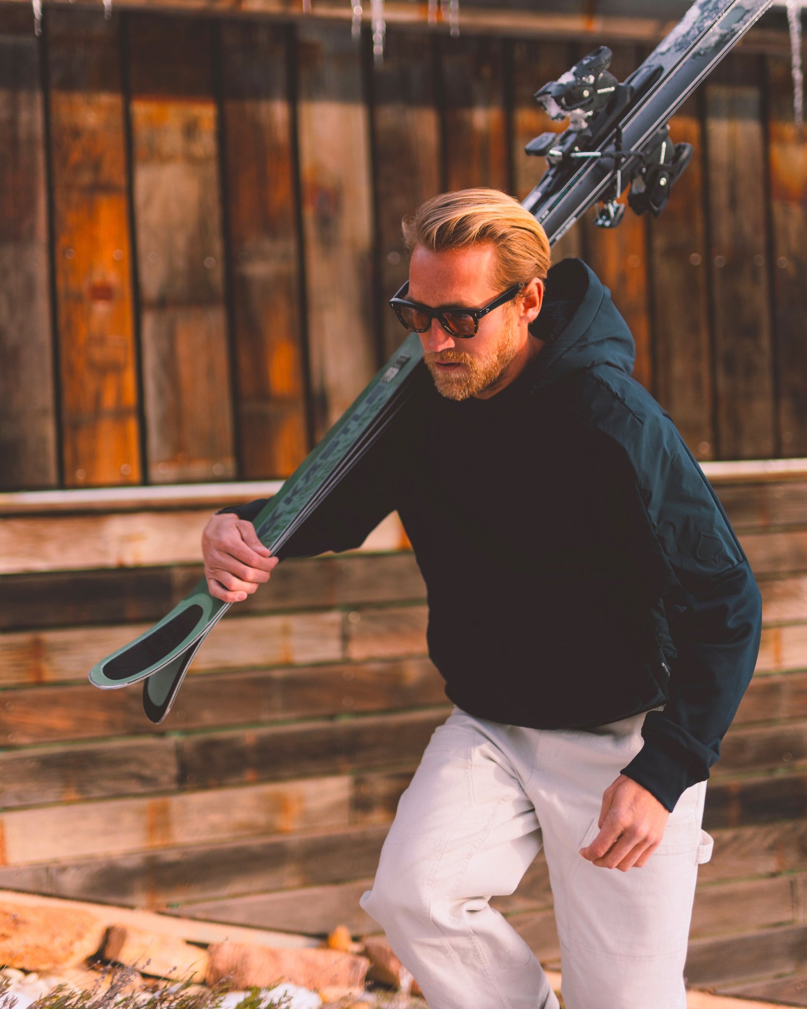 A person in an aspen Collection hoodie carries skis over their shoulder, walking past a rustic wooden building in the snow.