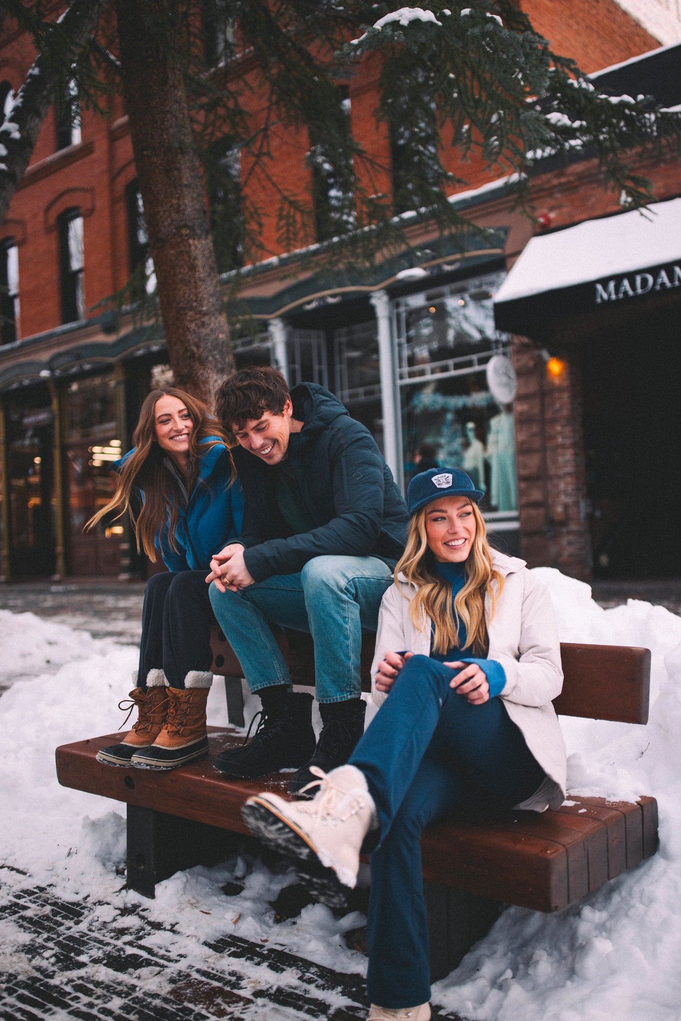 Three people sit on a bench, smiling in Aspen Collection new winter clothes on a snowy street with brick buildings.