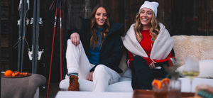 Two women smile on a sofa in Aspen Collection's winter attire, with ski gear and oranges nearby, framed by rustic wooden walls.