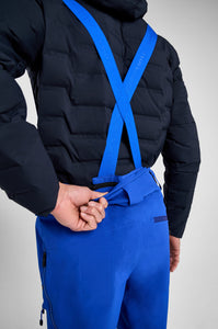 Person wearing a black puffer jacket and blue pants with suspenders on a gray background