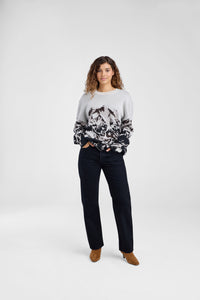 Woman wearing a sweater with black abstract mountain pattern and black jeans on a plain background