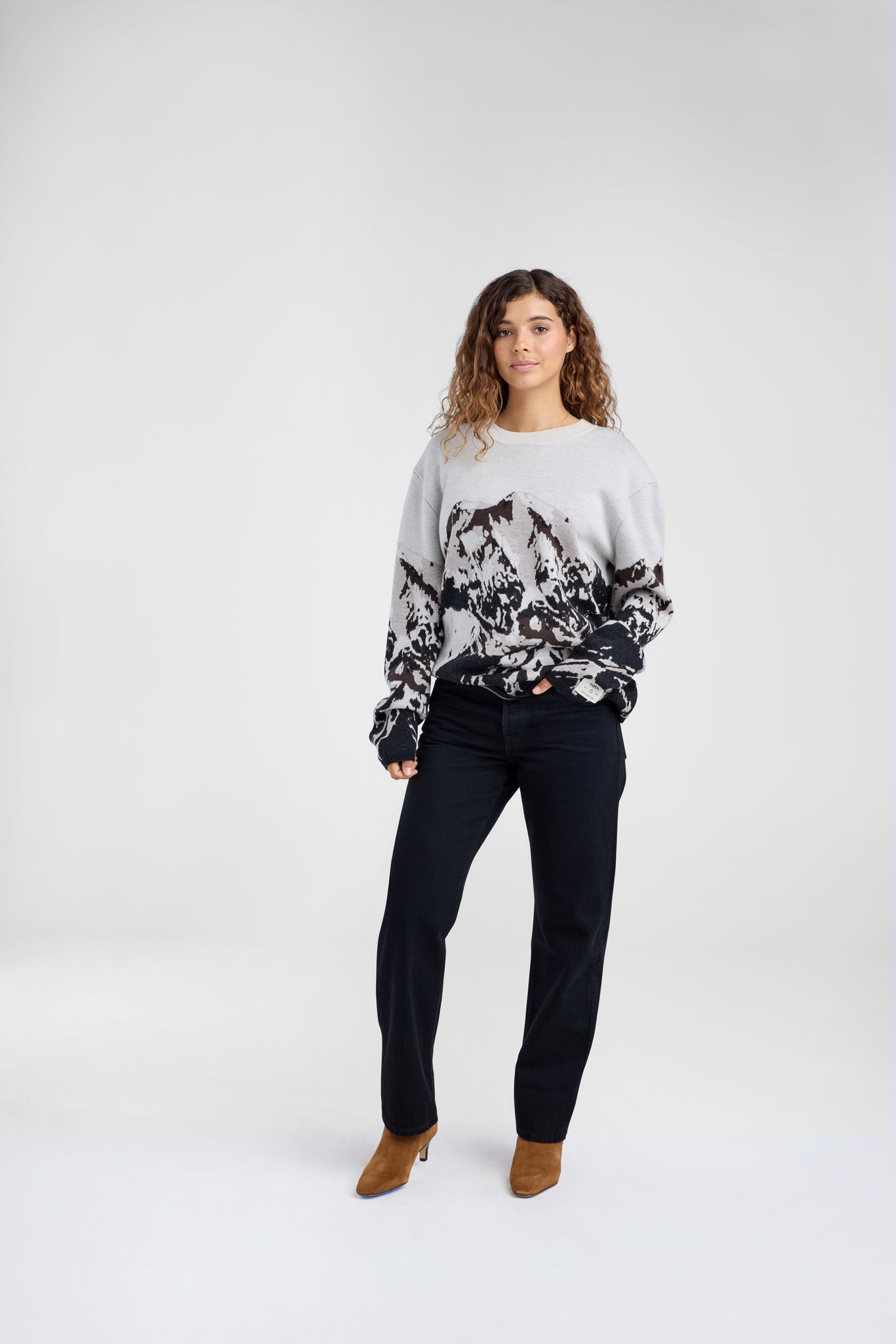 Woman wearing a sweater with black abstract mountain pattern and black jeans on a plain background