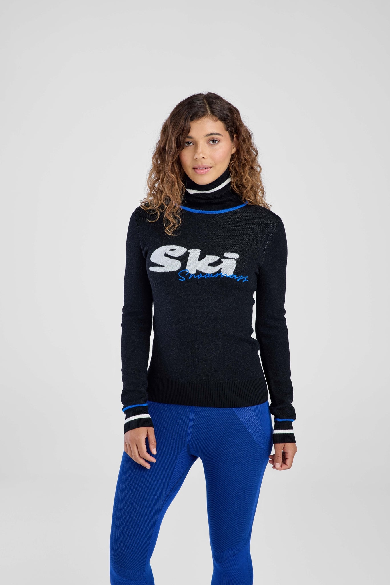 Woman wearing a black turtleneck with 'Ski' logo and blue pants on a white background