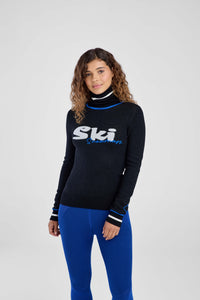 Woman wearing a black turtleneck with 'Ski' logo and blue pants on a white background