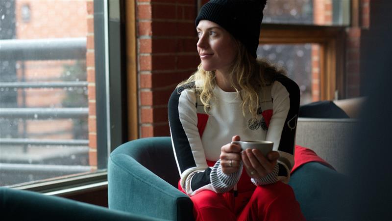 Woman sitting on a couch holding a mug, wearing a black beanie and red sweater with white patterns.