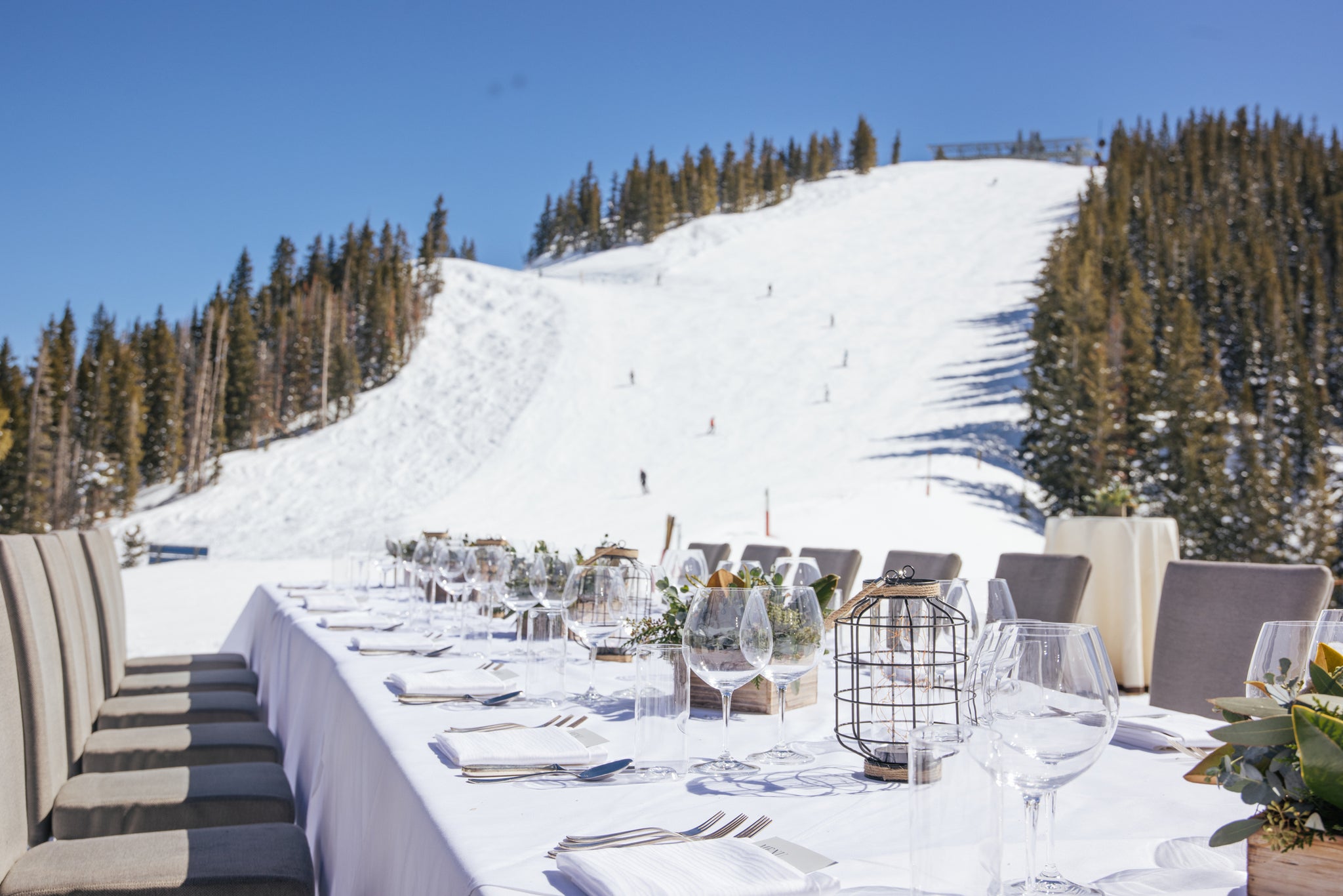 A long, elegantly set dining table with white tablecloths and glassware sits outdoors on a snowy mountain, surrounded by evergreen trees under a blue sky.