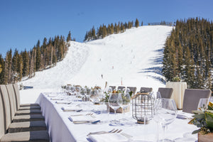 A long, elegantly set dining table with white tablecloths and glassware sits outdoors on a snowy mountain, surrounded by evergreen trees under a blue sky.