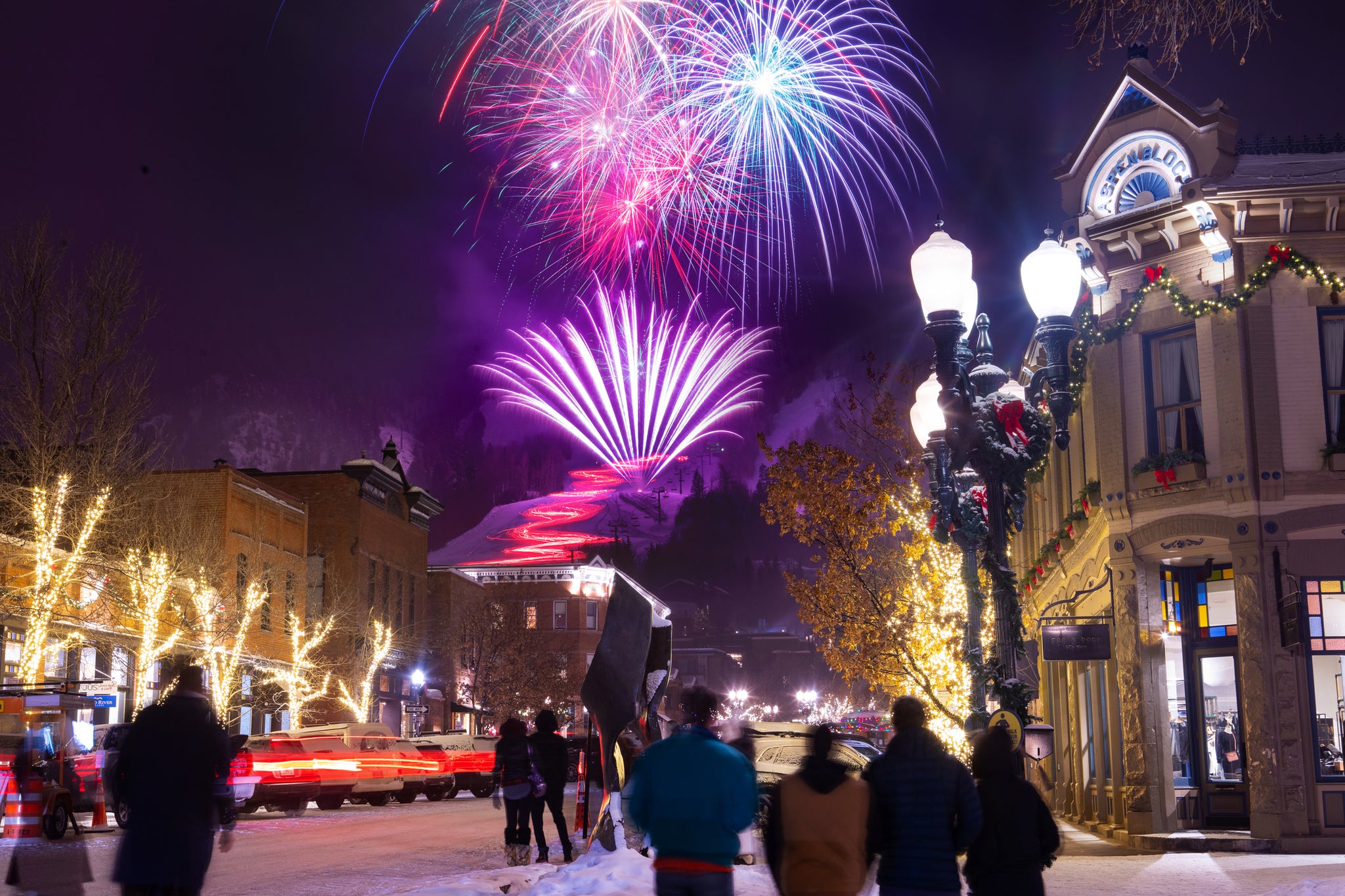 Fireworks burst over a snowy street lined with historic buildings. People watch amid holiday decorations and illuminated trees enhancing the festive scene in Aspen.